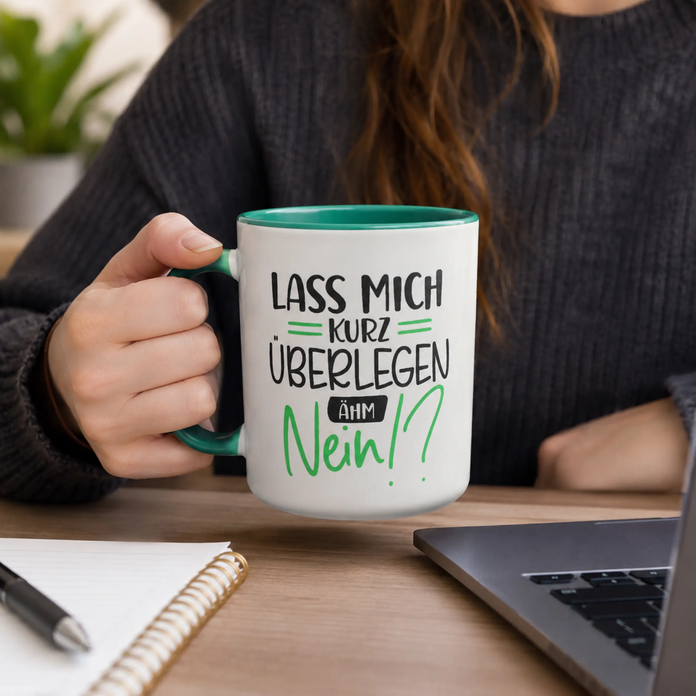 Tasse lass mich kurz überlegen mit grünem Henkel und Spruch Weiße Tasse mit grüner Innenseite und grünem Henkel, bedruckt mit dem Spruch „Lass mich kurz überlegen, ähm Nein!?“, in einer Hand am Schreibtisch gehalten.