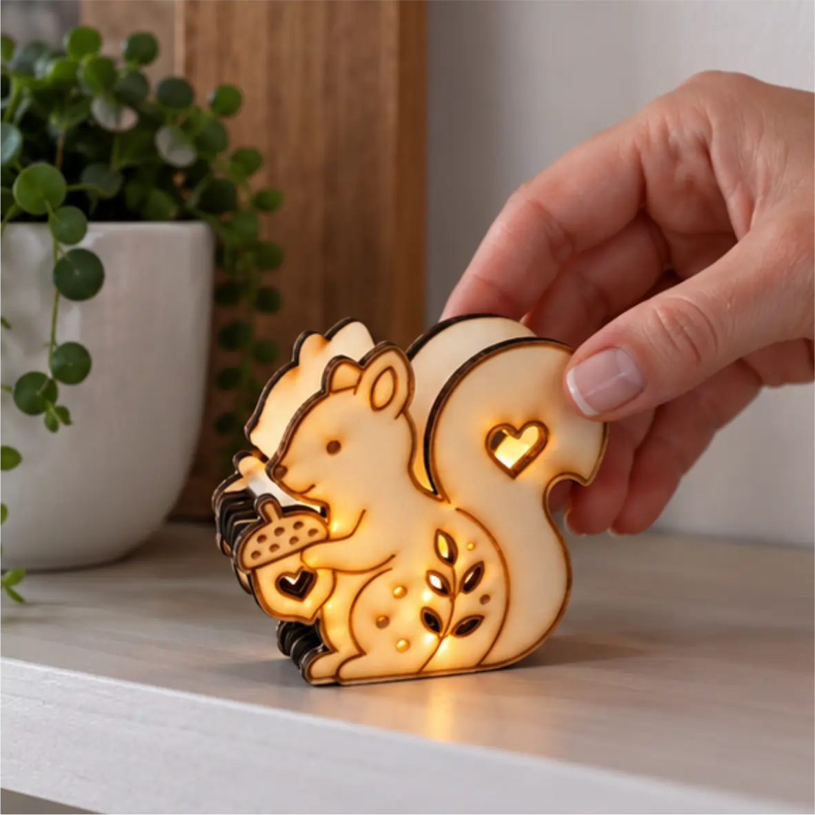 Warm leuchtendes LED-Eichhörnchen aus hellem Sperrholz mit Eichel, großem Schwanz sowie Herz- und Blattausschnitten. Eine Hand hält die Figur auf einer hellen Ablage neben einer Topfpflanze.