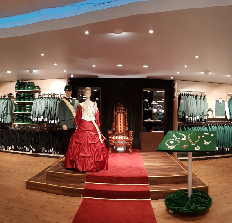 Ausstellungsfläche mit Schaufensterpuppen in Uniform und rotem Ballkleid Ausstellungsfläche in einem Bekleidungsgeschäft mit zwei Schaufensterpuppen, eine in grüner Uniform und eine in rotem Ballkleid mit Schärpe, dazu ein verzierter Thron, rote Teppichstufen, grüne Kleidung an den Wänden und Schmuck auf einem Tisch.