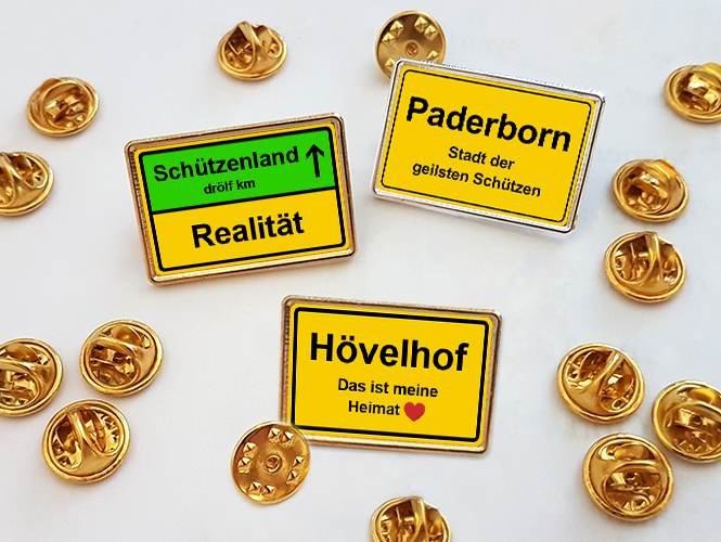 Ansteckpins mit Ortsschildern Paderborn, Hövelhof und Schützenland Drei rechteckige Ansteckpins im Ortsschild-Design mit den Aufschriften Paderborn, Hövelhof und Schützenland auf weißem Hintergrund, umgeben von goldfarbenen Pin-Verschlüssen.