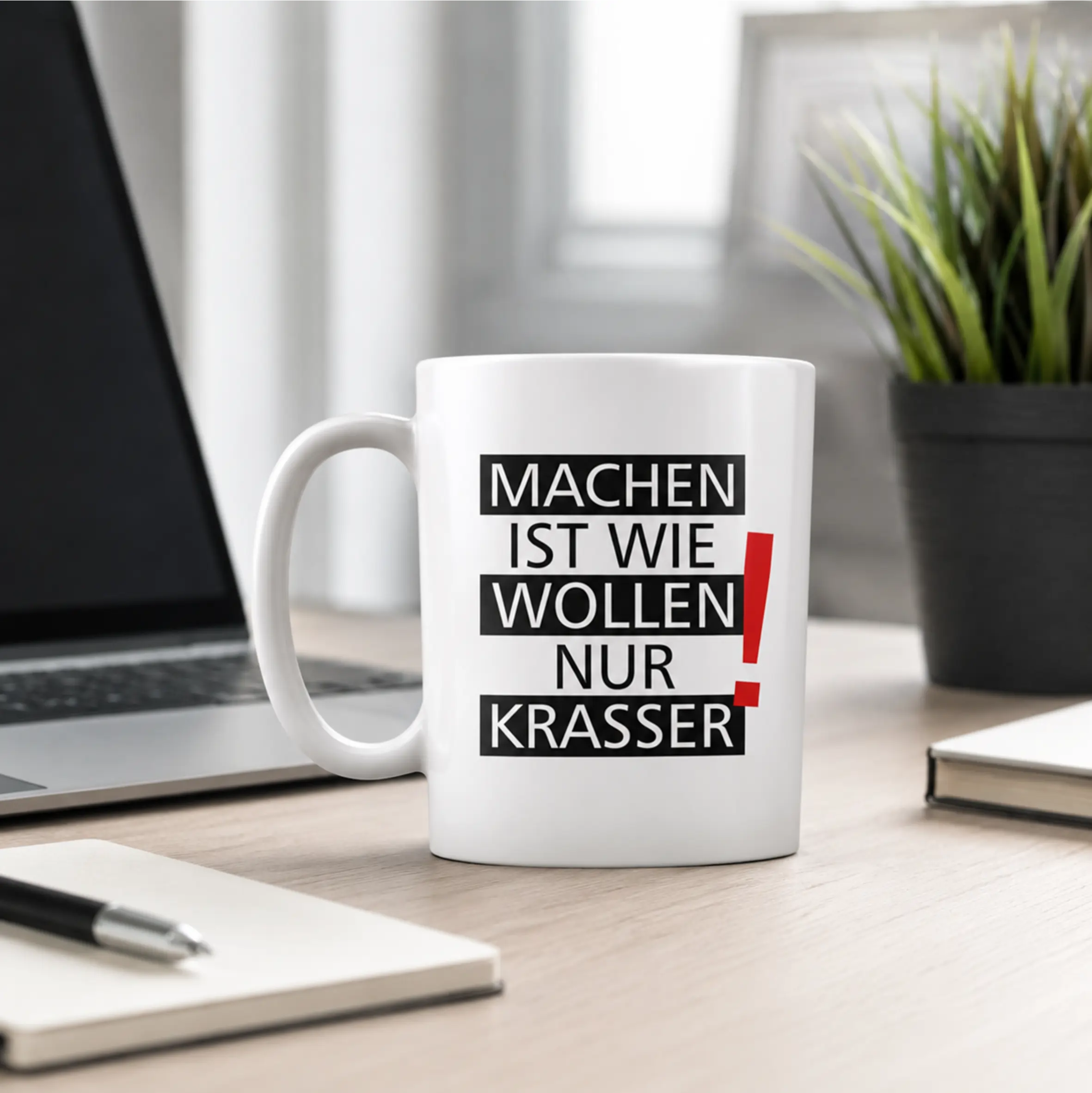 Weiße Keramiktasse mit C-Henkel auf einem Schreibtisch, bedruckt mit dem Spruch „Machen ist wie wollen nur krasser“ in schwarzer Typografie und rotem Ausrufezeichen.