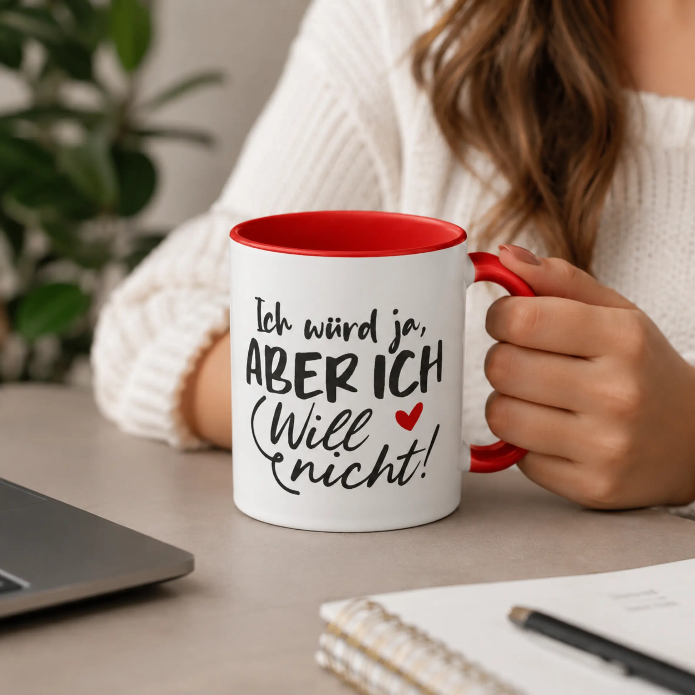 Tasse Ich würd ja aber ich will nicht mit rotem Henkel Weiße Tasse mit roter Innenseite und rotem Henkel, bedruckt mit dem Spruch „Ich würd ja, aber ich will nicht!“, in einer Hand gehalten auf einem Tisch.