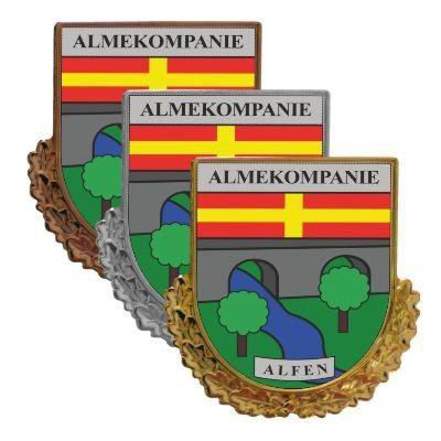 ALMEKOMPANIE ALFEN Abzeichen mit Wappenschild Drei überlappende Abzeichen mit Wappenschild, Schriftzug ALMEKOMPANIE und ALFEN, rot-gelb gestreiftem Balken mit Kreuz sowie Landschaftsmotiv in Grün und Blau, jeweils mit bronzefarbenem, silberfarbenem und goldfarbenem Rand