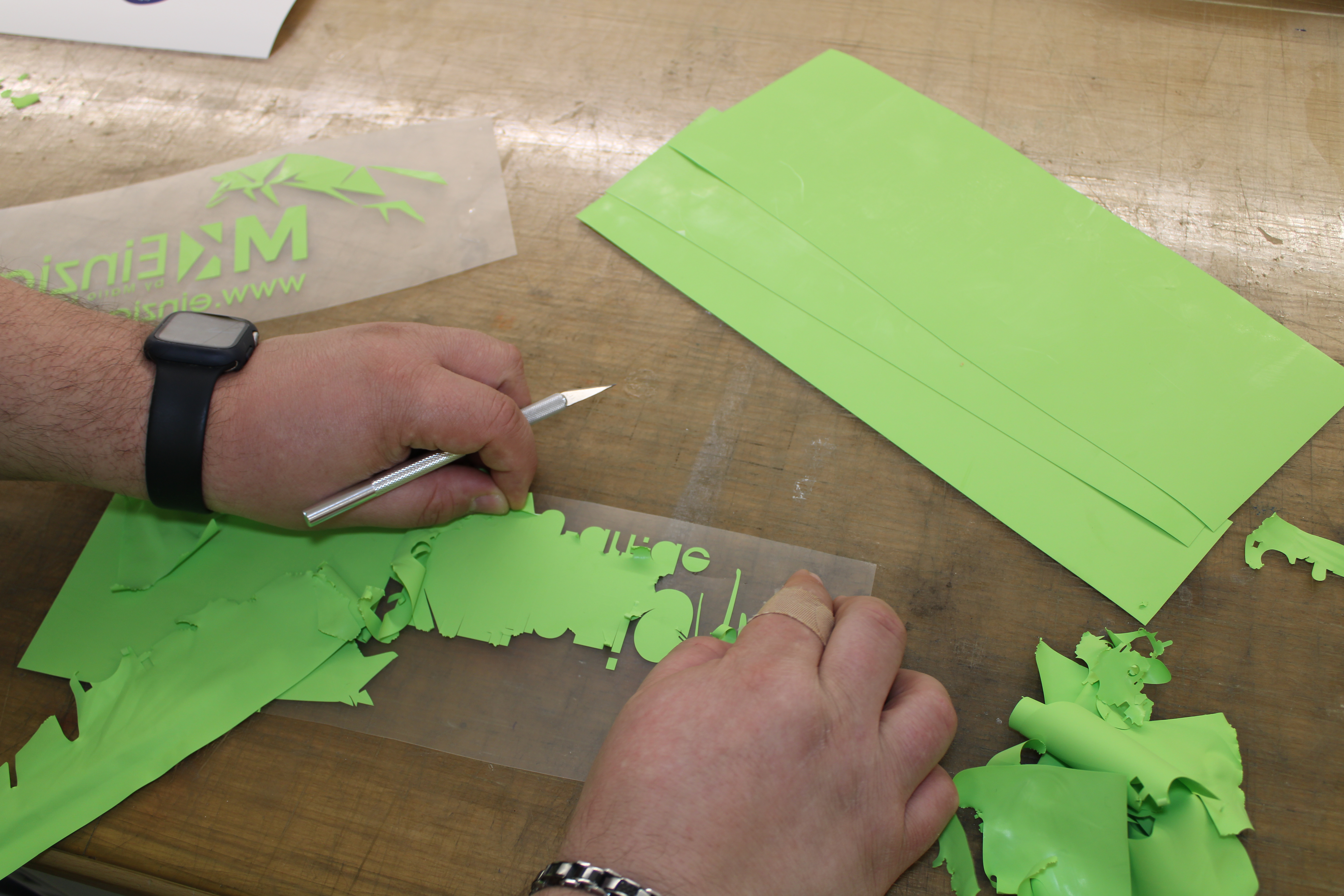 Grüne Schriftzüge und Motive auf transparenter Trägerfolie werden von Hand mit einem Präzisionsmesser entgittert. Auf der Arbeitsfläche liegen grüne Bögen und entfernte Folienreste.