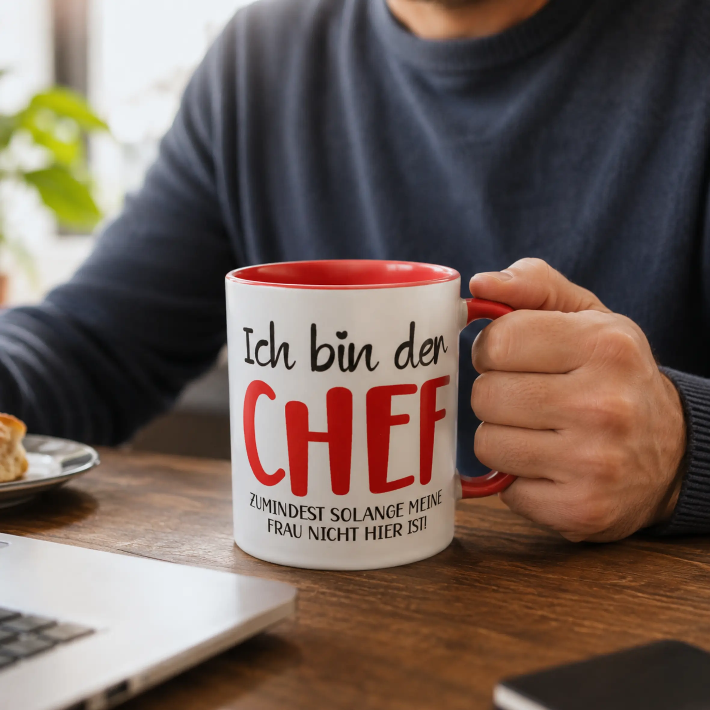 Weiße TwoTone-Tasse mit roter Innenseite und rotem Henkel, bedruckt mit dem Spruch „Ich bin der Chef, zumindest solange meine Frau nicht hier ist“, auf einem Tisch in einer Hand gehalten.
